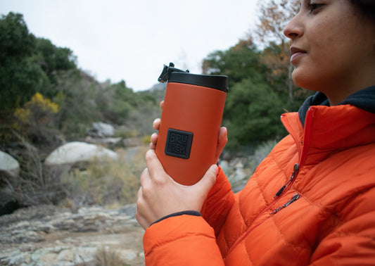 Person holding an orange French press coffee maker outdoors with a natural background
