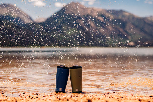 Pair of coffee tumblers on a sandy beach with water splashing around them and mountains in the background.