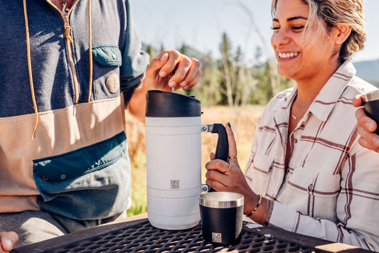Two people enjoying making coffee outdoors at a picnic table with a scenic background.