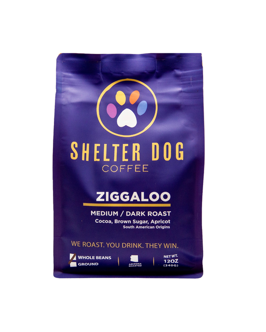 A 12 oz bag of Shelter Dog Coffee Ziggaloo medium/dark roast whole beans, featuring a deep purple bag with gold lettering and a colorful paw print logo, set against a plain white background.