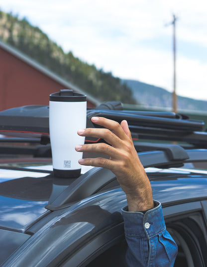 Person holding a white insulated tumbler mug with a car roof rack in the background