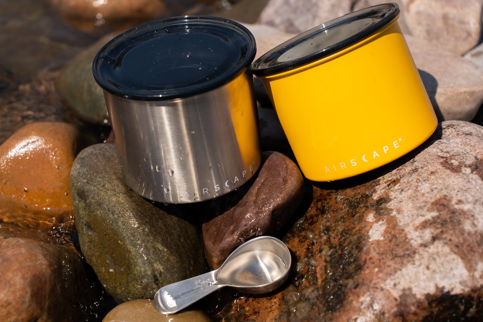 Two metal containers with lids on rocks, one yellow and one silver, with a spoon nearby.