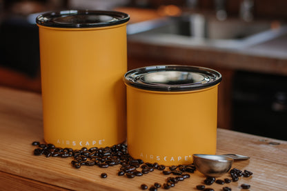 Two yellow Aircscape coffee containers on a wooden surface with coffee beans and a scoop.