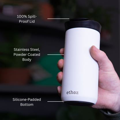 White travel mug with black lid held by a hand, labeled features on a blurred background