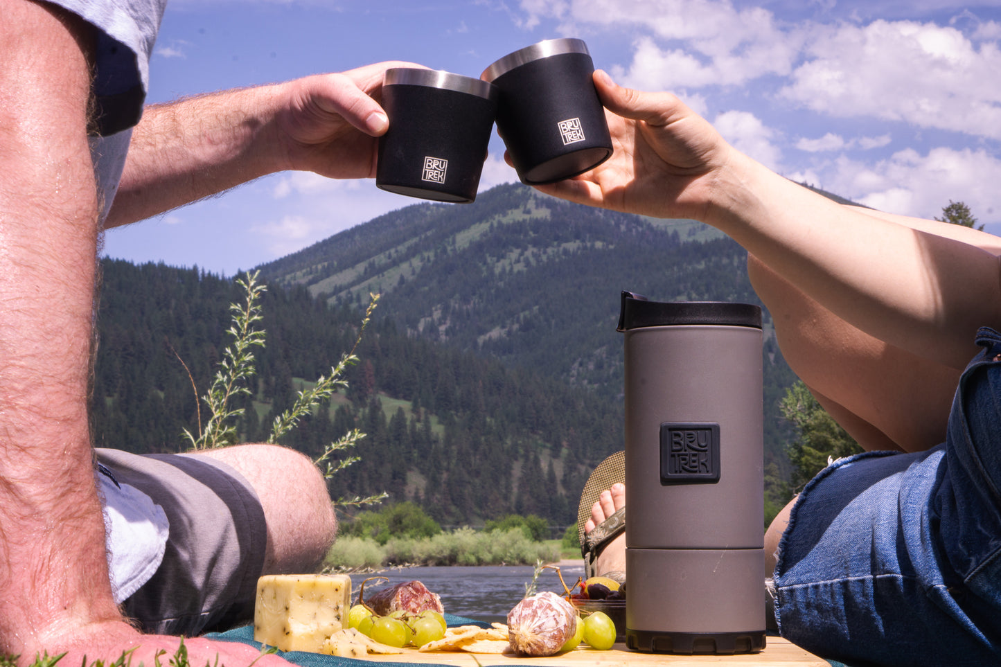 Two people toasting with black travel mugs next to a gray travel French press coffee maker while outdoors near a lake with mountains in the background.