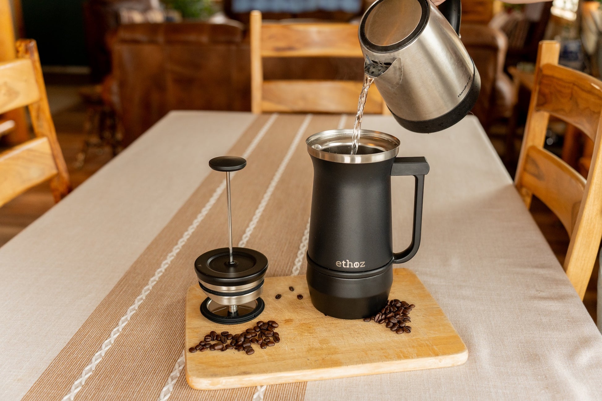 Black French Press coffee maker on a wooden board with coffee beans and a kettle pouring water, set against a restaurant interior.